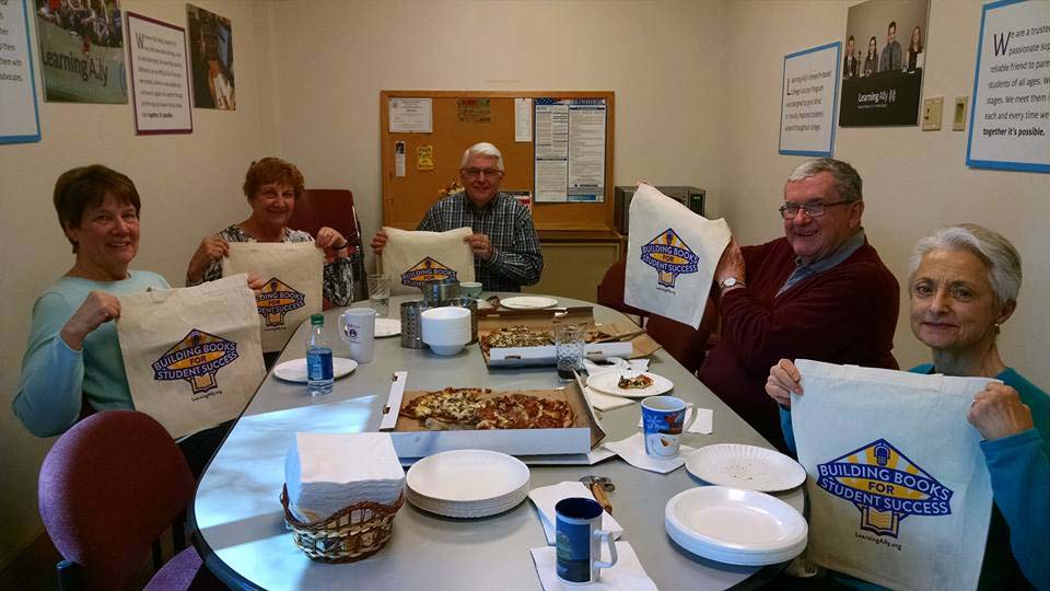 Jim and volunteers holding up Building Books fundraiser towels and sitting down for pizza in Orland Park Studio